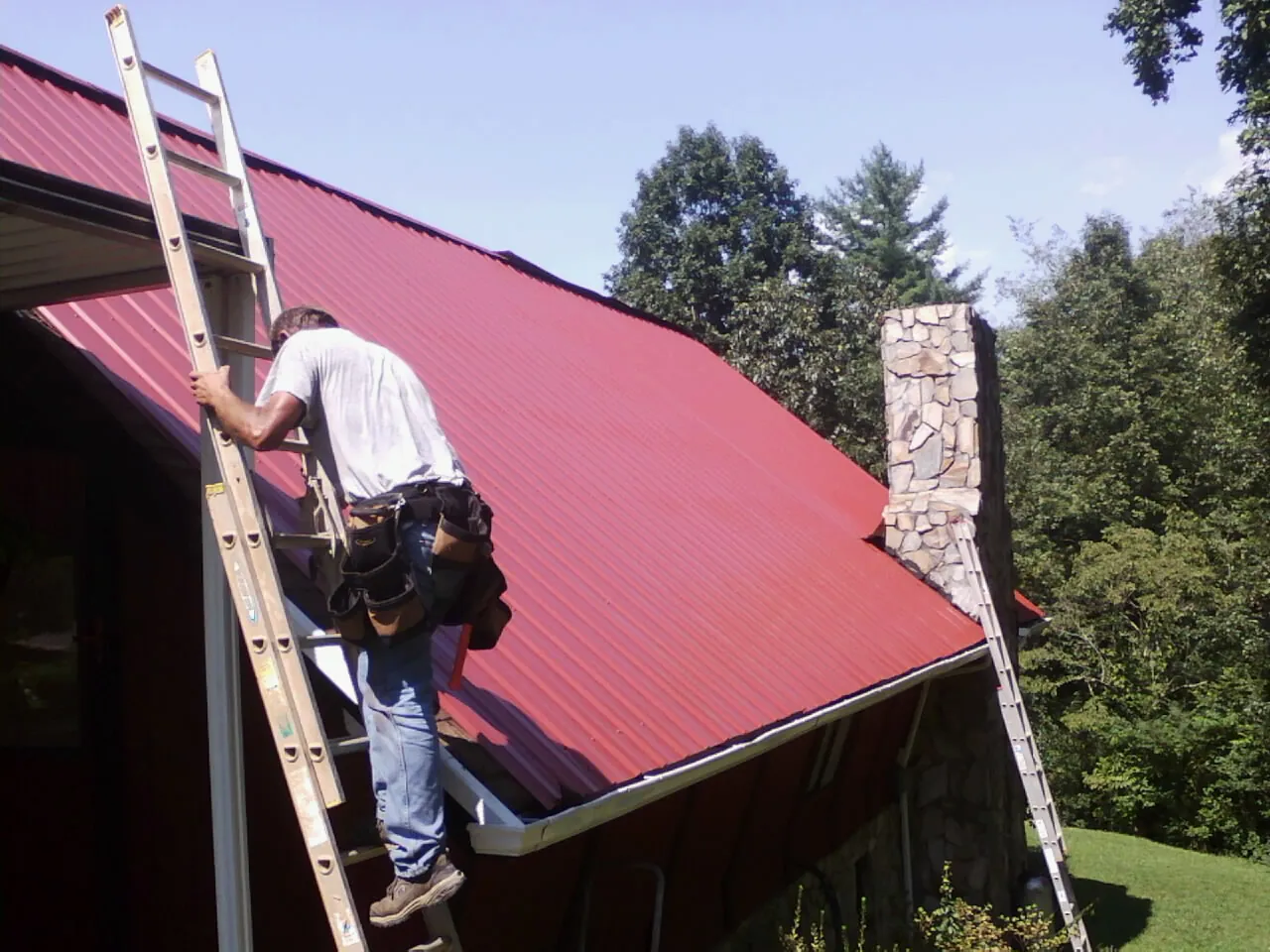 Roofer on a ladder beside a red metal roof and stone chimney — Soffit & Fascia Repair by Roofing Brothers in Guffey